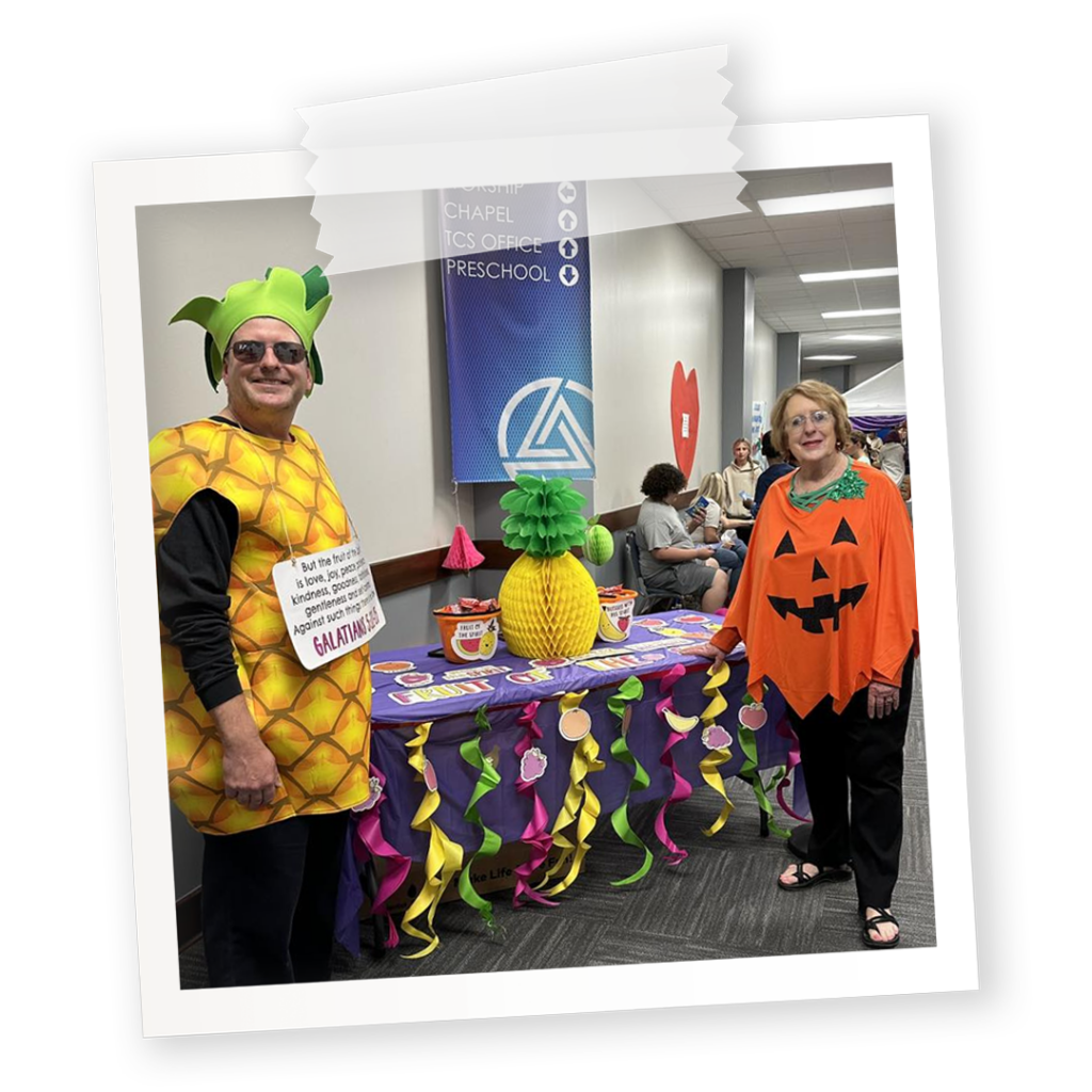 A church trunk-or-treat booth for the fruits of the spirit. Features man as a pineapple and woman as a pumpkin in front of a colorful booth decorated with fruit