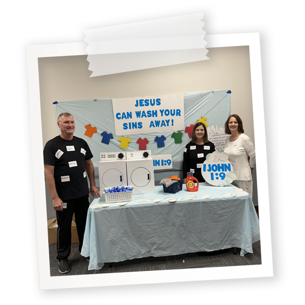 A church trunk-or-treat booth for John 1:9. Jesus can wash away your sins. Three people standing in front of the booth that has a washing machine and laundry detergent and bubbles.